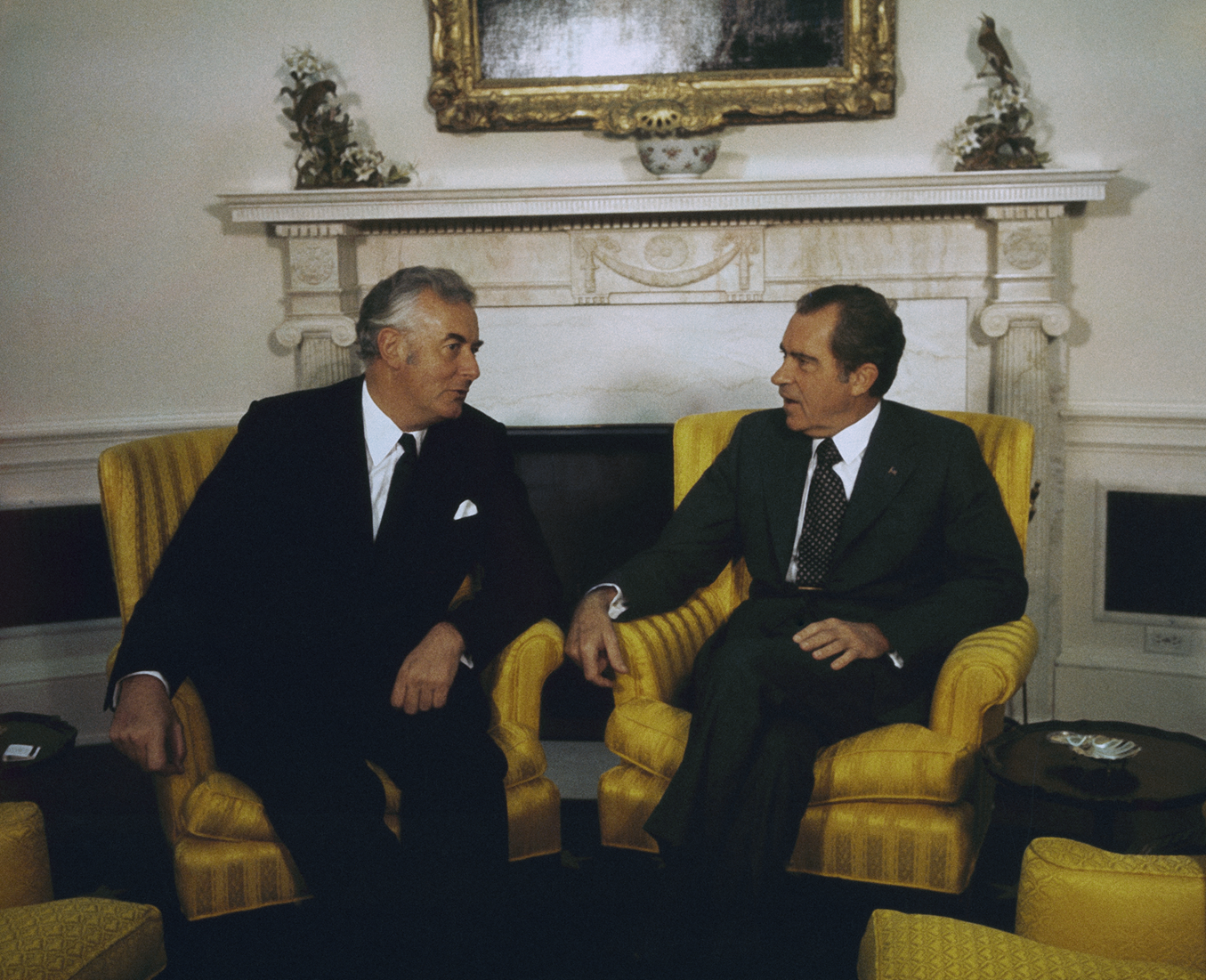 Richard Nixon and Gough Whitlam sit in yellow chairs in the Oval Office facing each other and talking.