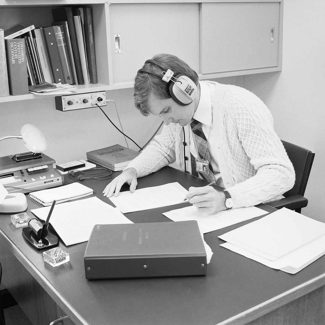 A man with headphones on is writing on a sheet of paper. He is in an office surrounded by papers.