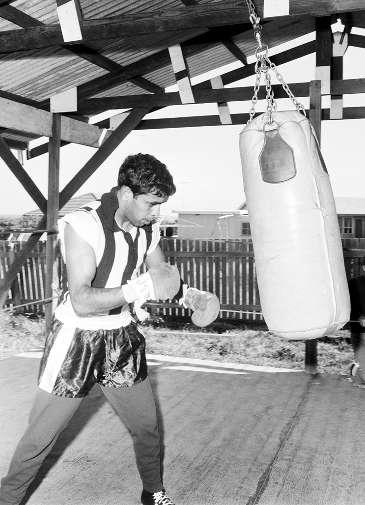 Lionel Rose is dressed in boxing gear. He is looking at a punching bag. 