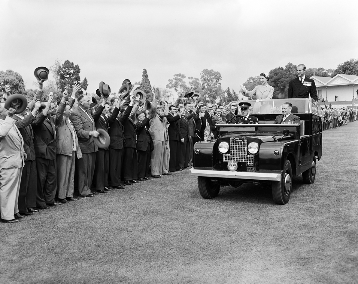 The Queen and Prince Phillip are standing in the back of a car waving to a crowd who are all waving their hats to them.