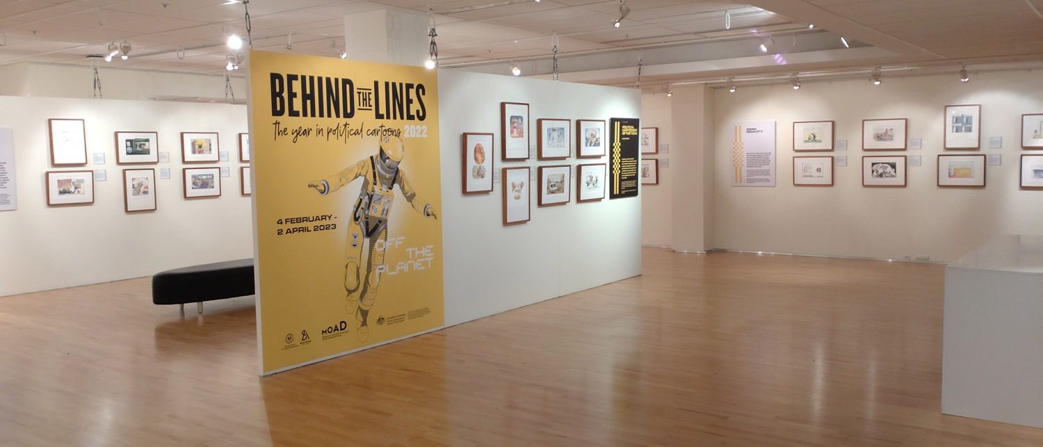 A gallery space with framed cartoons on the walls. A large, yellow introductory panel features the illustration of an astronaut and the words 'Behind the Lines'.