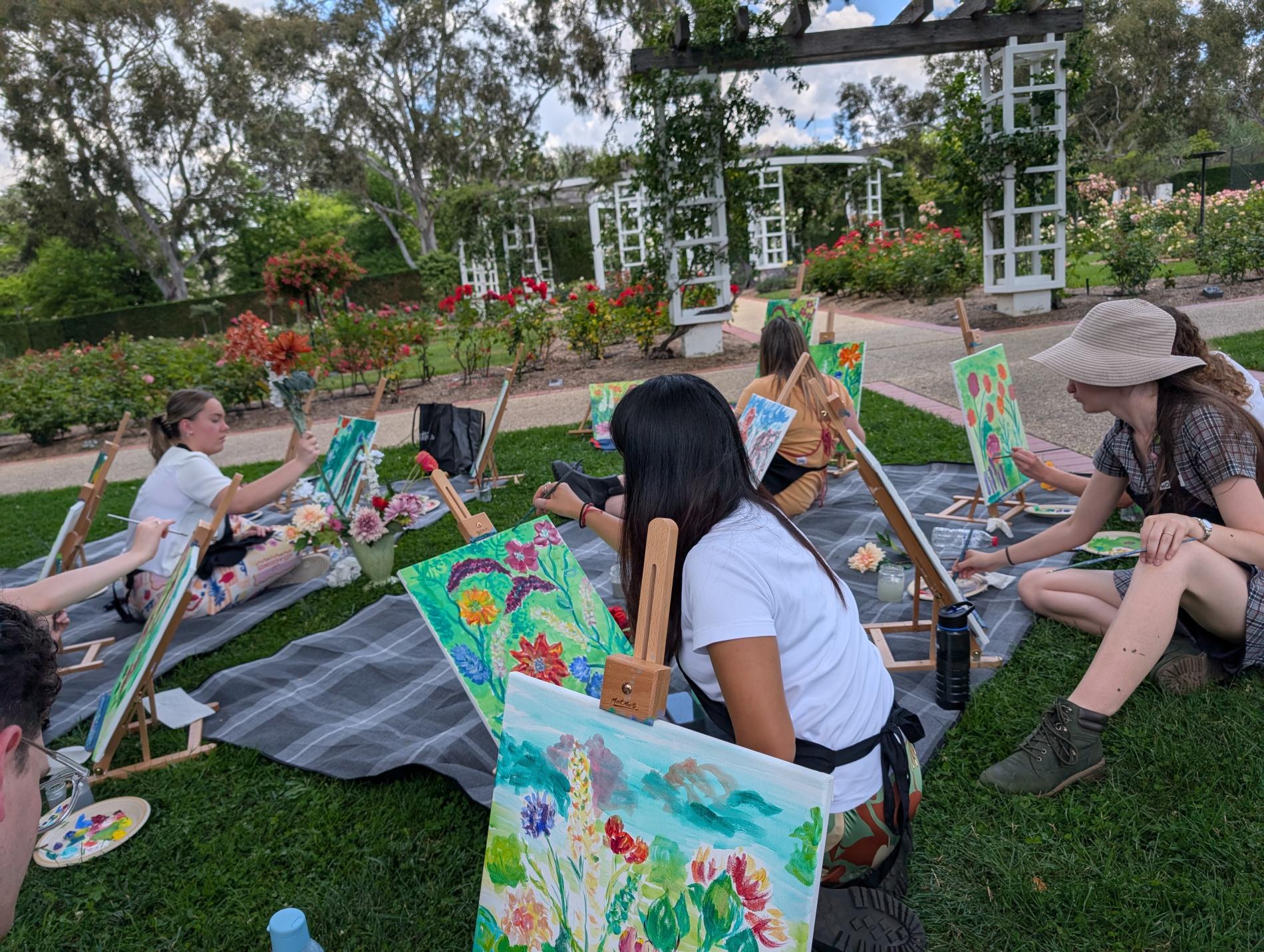 People sit on the grass in the rose gardens at Old Parliament House painting floral artworks.