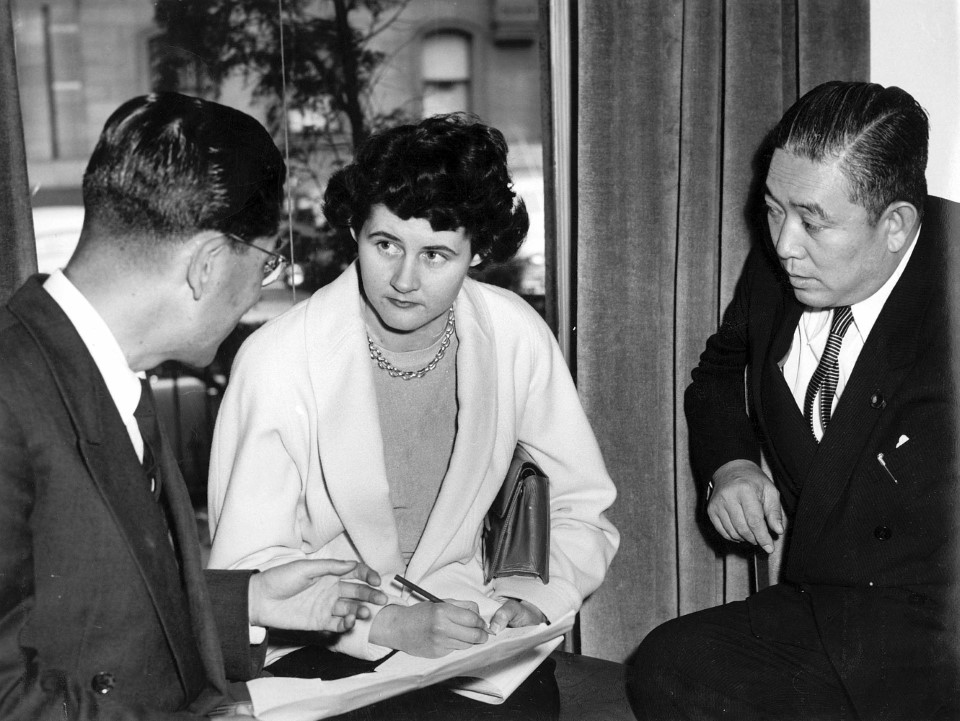Reporter Patricia Clarke interviewing two Singaporean diplomatic delegates. Taking notes in a notepad on her lap while talking to the two men.