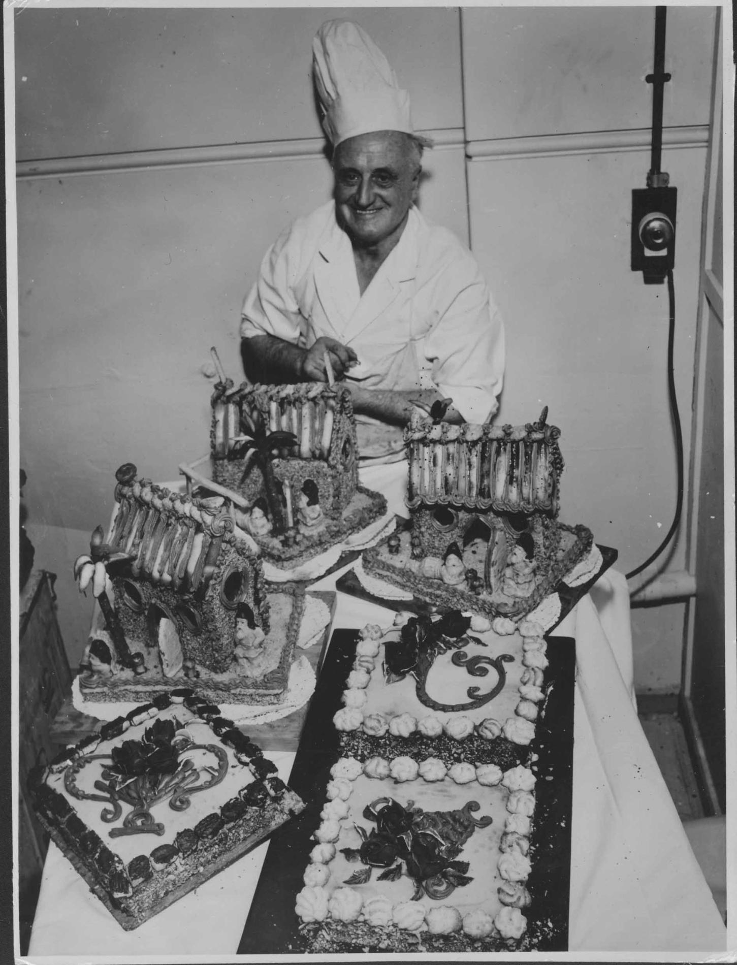 A black and white image of a chef standing behind a table filled with different desserts.