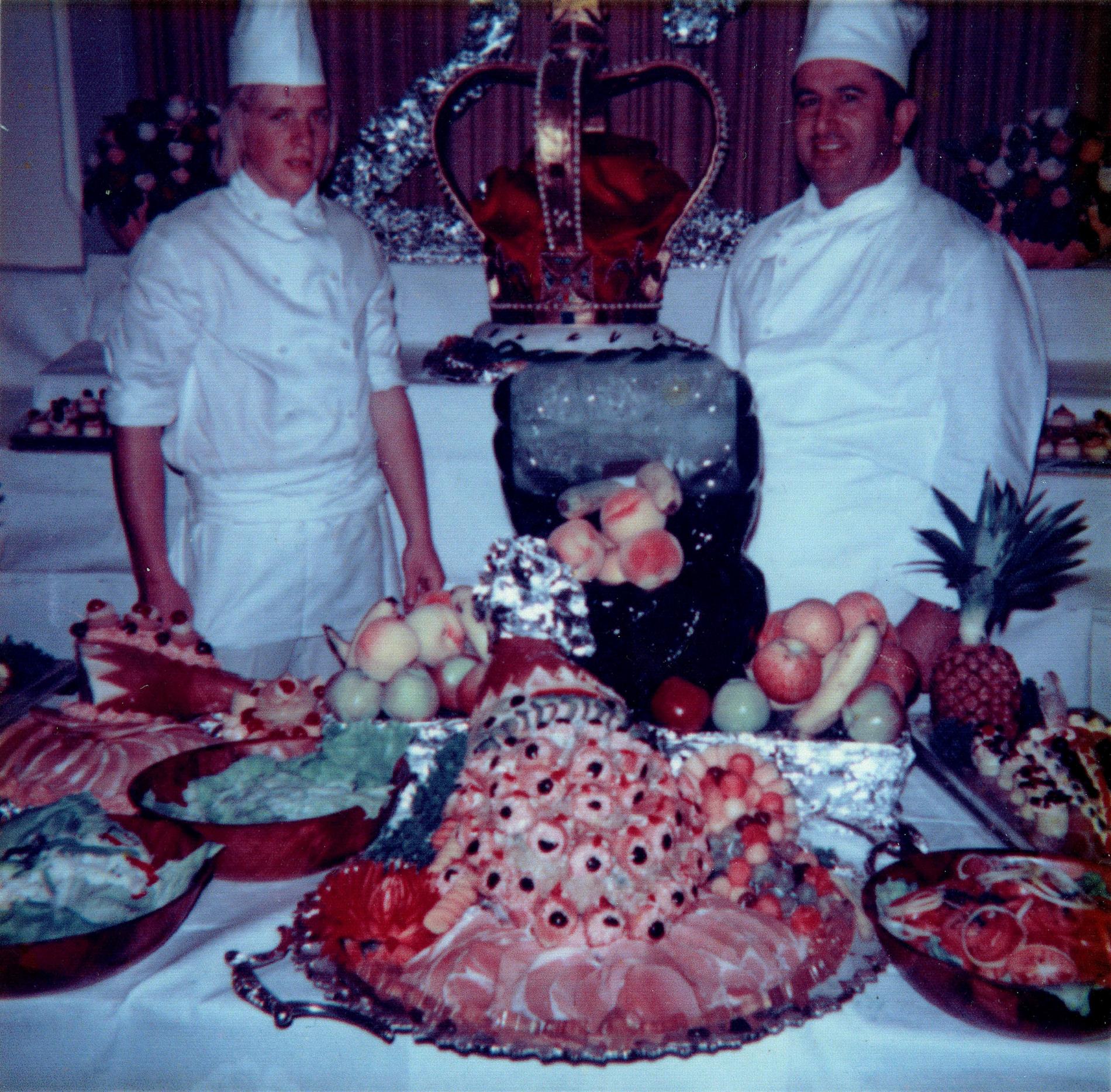 Two chefs standing behind a table covered in fruit and desserts.