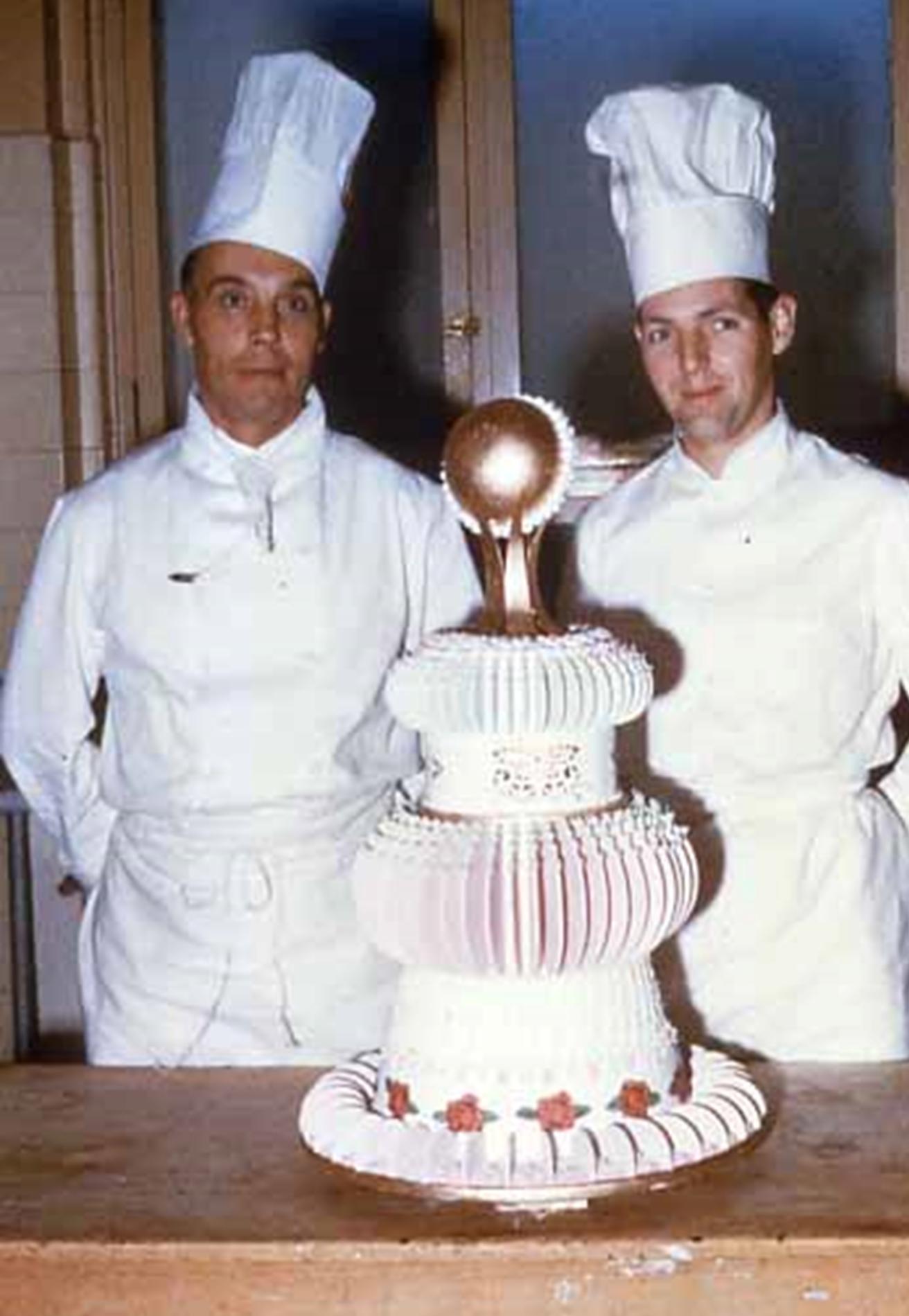 Two chefs, both wearing hats, standing next to a layered cake with a topper.