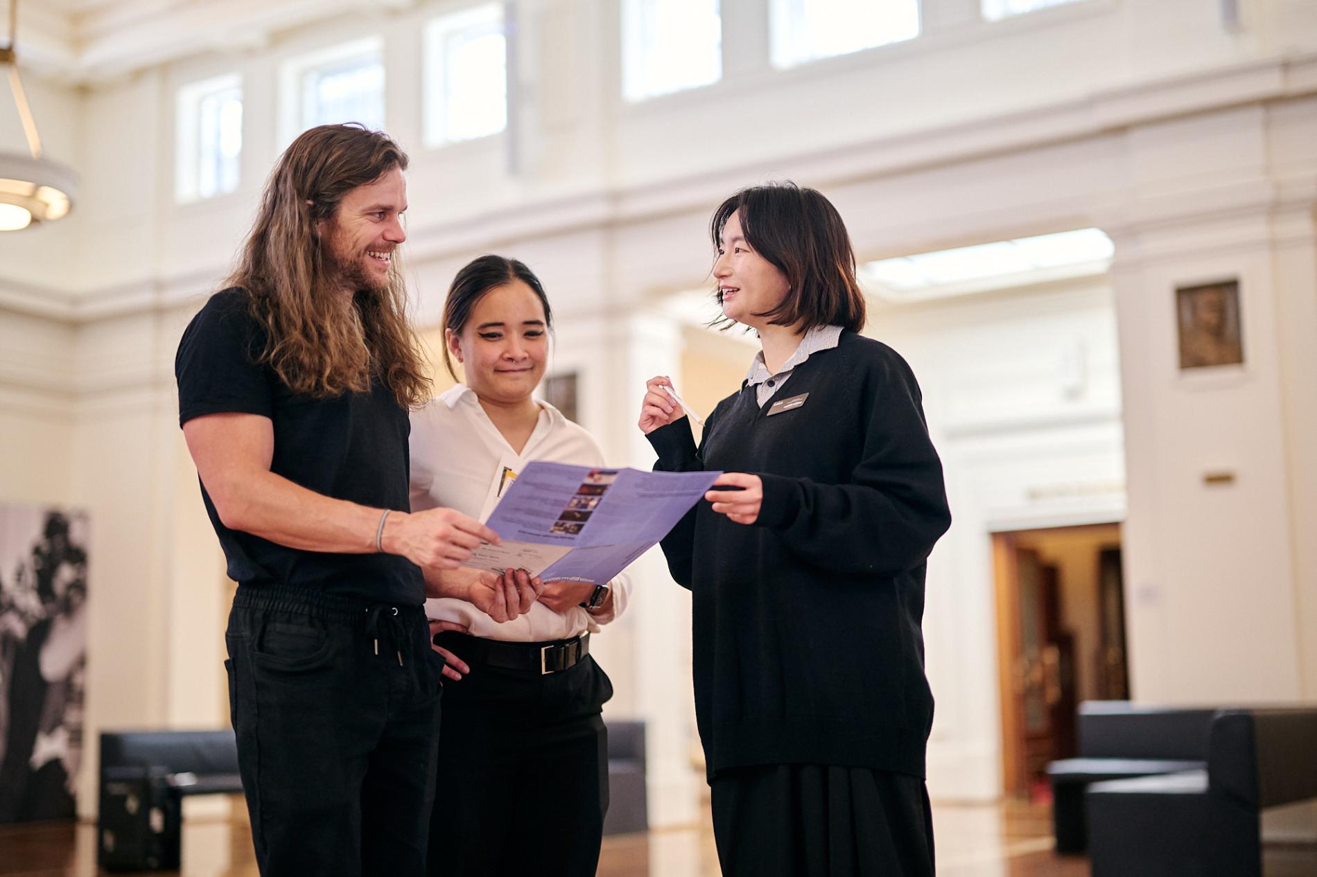 Three people gathered around a brochure in Kings Hall, Old Parliament House.