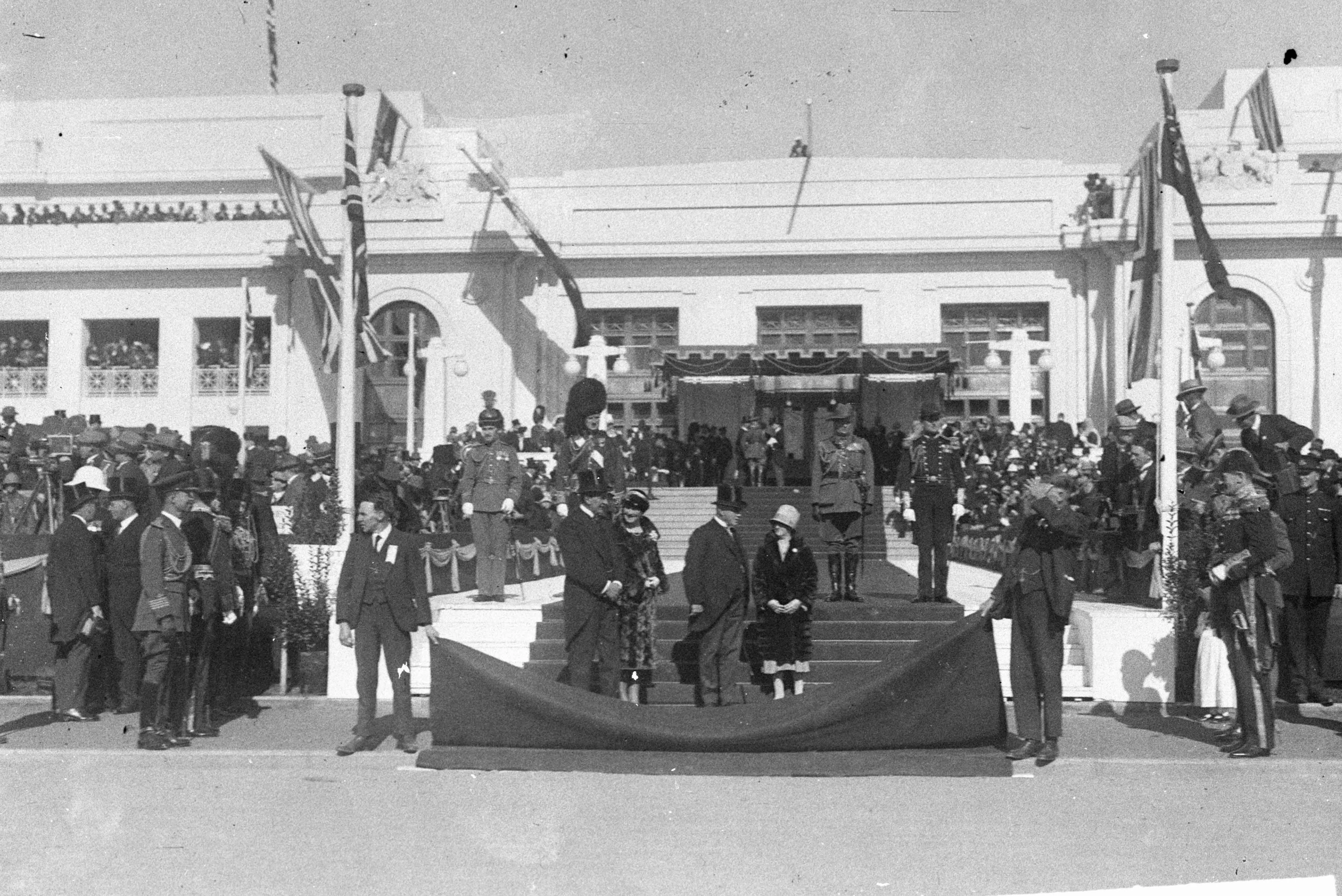 This black and white photograph captures the façade of Parliament House decorated with flags, a canopy and welcome carpet just before the arrival of the Duke and Duchess of York for the opening of the building. The weather is sunny and a bit breezy. There are expectant crowds on the roof, front steps, verandahs and flanking the front door. A guard of honour in military uniform stands at the ready. Two attendants are holding up the end of the red carpet to keep it clean until the Royal carriage arrives. We k