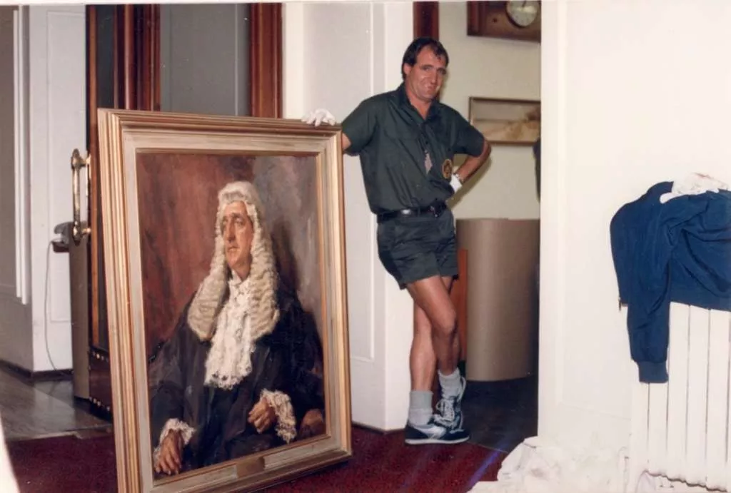 A removalist wearing white gloves posses next to a portrait painting of a main wearing the President of the Senate's wig, black gown and lacy collar. A removalist wearing white gloves posses next to a portrait painting of a main wearing the President of the Senate's wig, black gown and lacy collar.