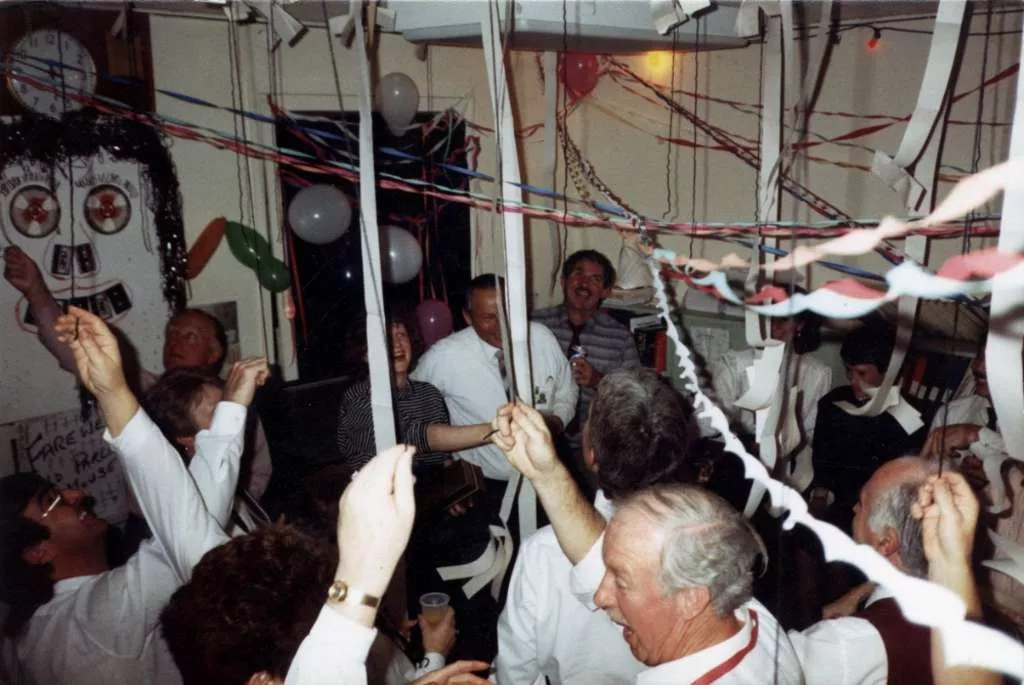 A crowded office with streamers decorated across the room and people celebrating with drinks.