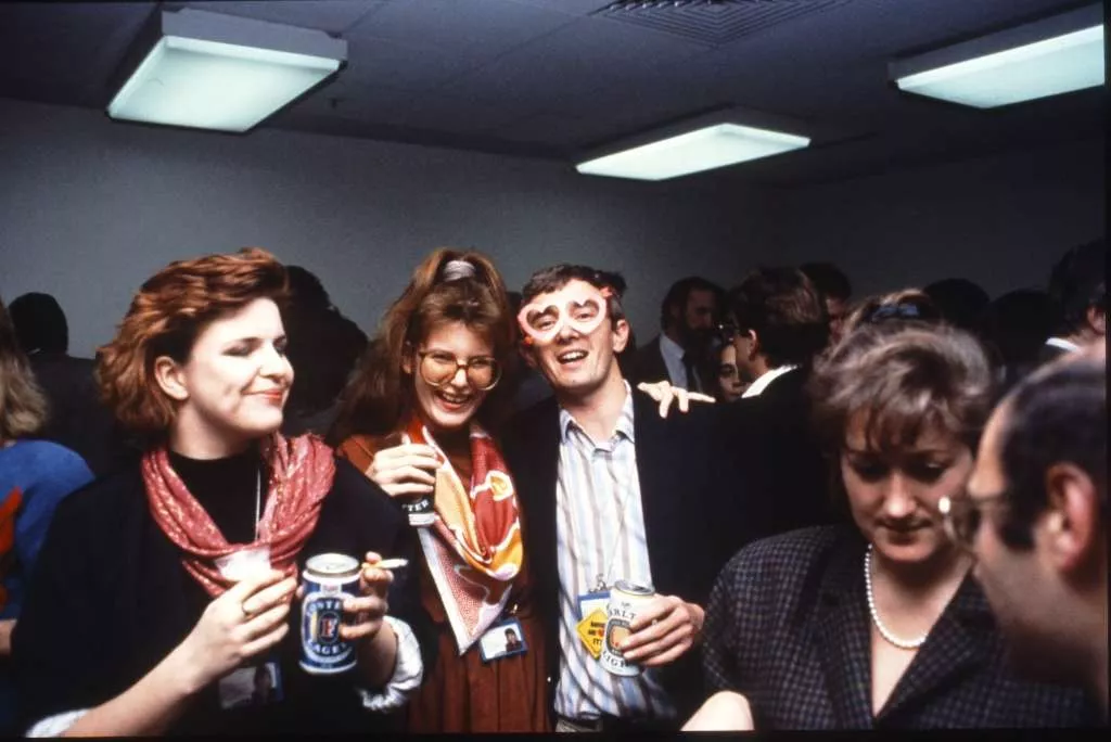 Office colleagues celebrating with beers and cigarettes in a crowded room.