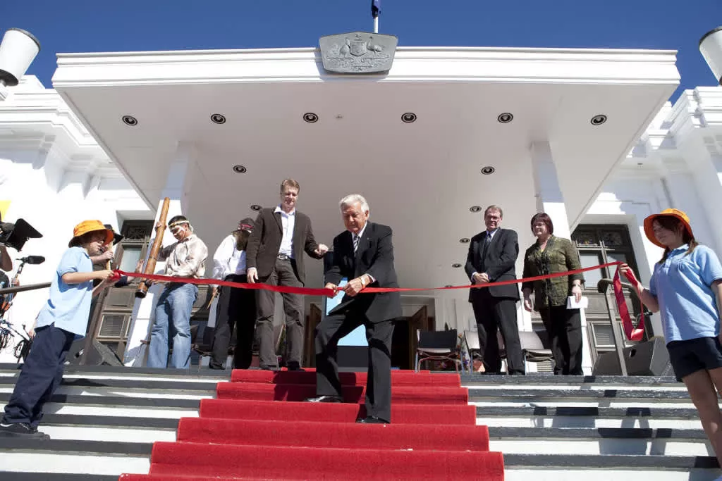 Bob Hawke stands on a red carpet at the front steps of Old Parliament House, and cuts a red ribbon held by two children.