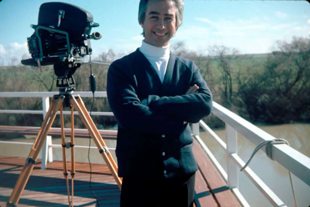 A man, Don Dunstan, stands on decking overlooking a river, wearing a white skivvy and dark cardigan, with a film camera on a tripod in the background.