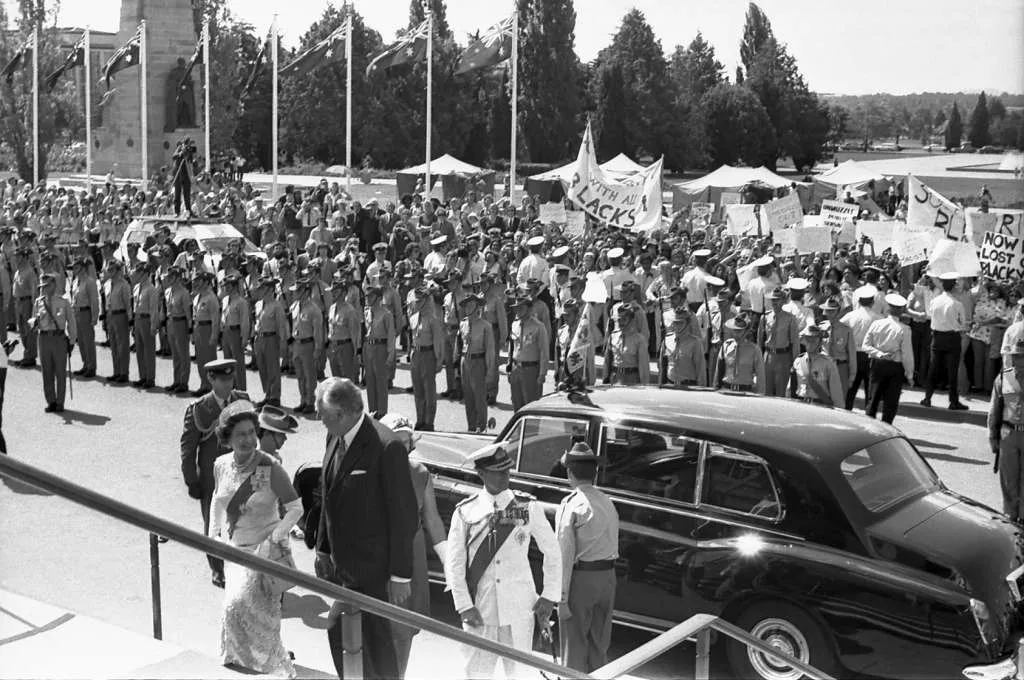 Queen Elizabeth walk up the steps to Parliament House with Gough Whitlam, attended by a parade of troops, and in the background a crowd protests with placards and flags.