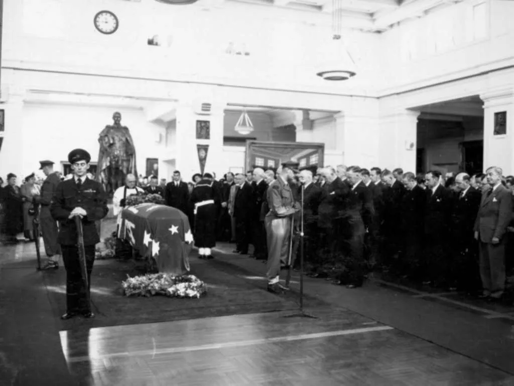 Four armed servicemen stand around a coffin draped in the Australian Flag, whilst a large group of men stand in rows, their heads bowed.