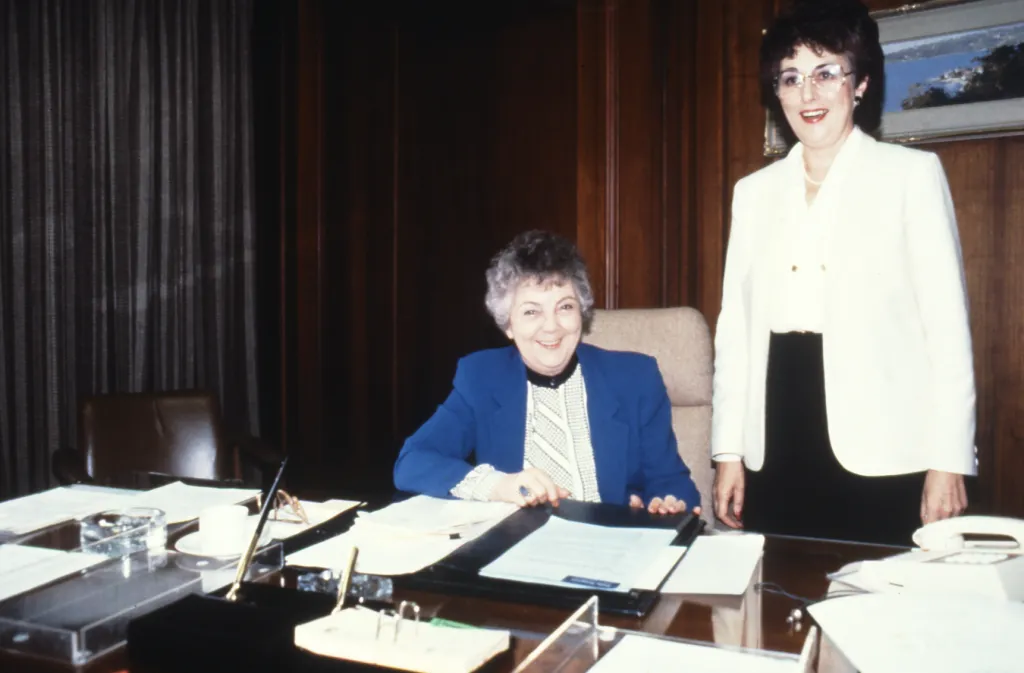 Speaker Joan Child and Marie Donnelly standing behind her desk. 1988, film photo. 