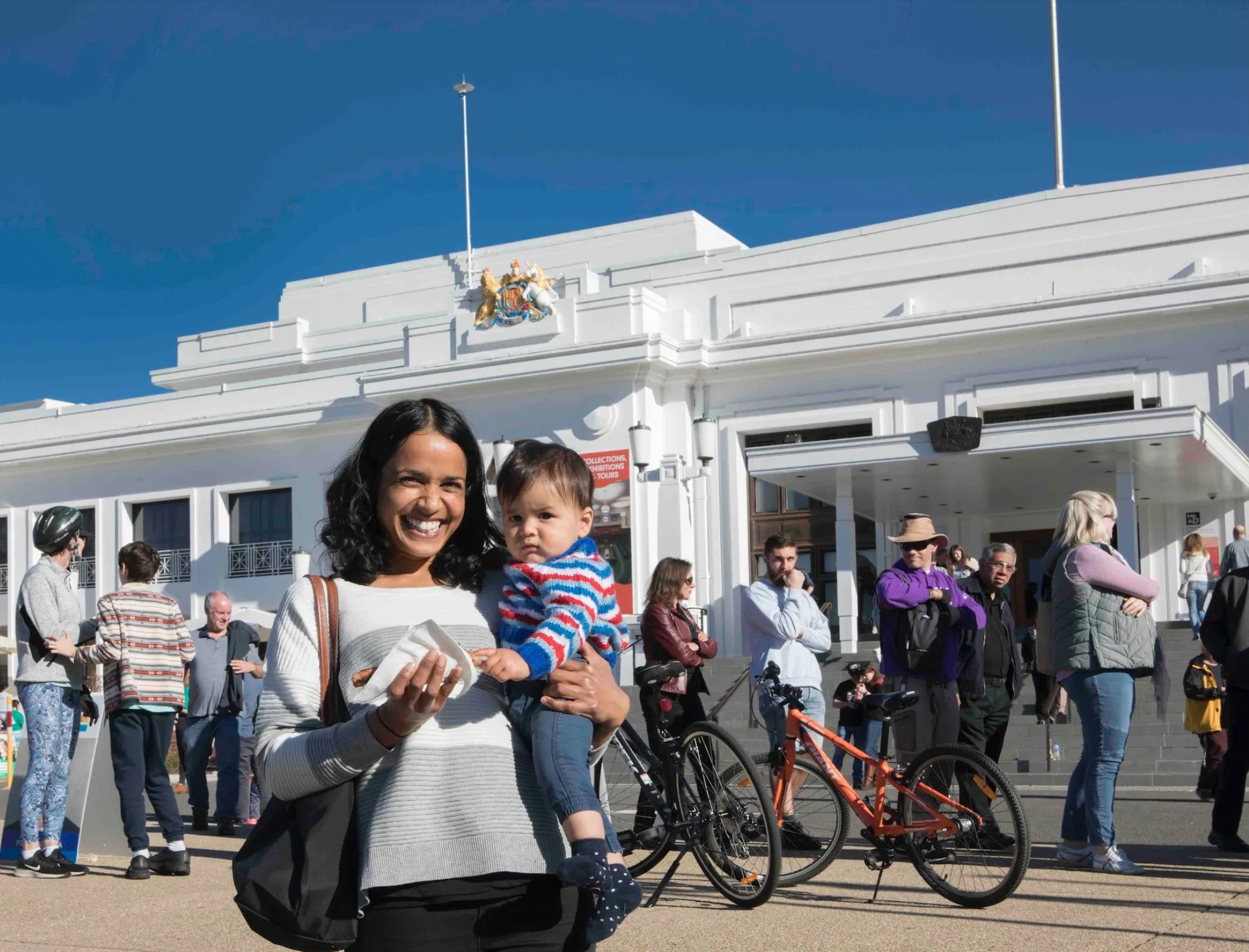 A mother holds her baby and smiles at the camera at the front of Old Parliament House. A row of people waiting to vote are behind her. She holds a sausage sandwich.