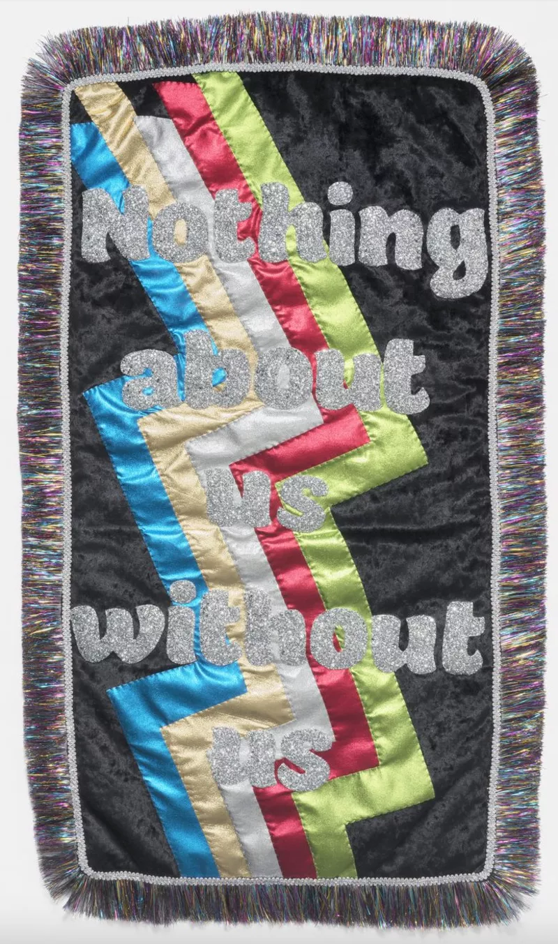 Embroidered, multicoloured banner with black sides and black tassels around it, that says 'Nothing about us without us'. 