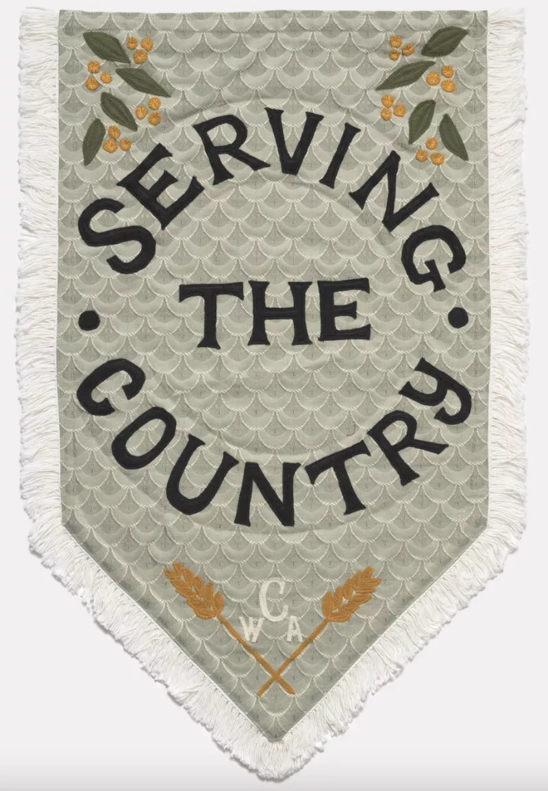 Soft green coloured tapestry with wattle and leaf motif on the left and right top corners. Underneath are the words 'Serving the country' and below that the acronym for the Country Women's Association, 'CWA'.