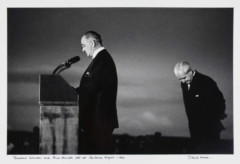 President Lyndon Johnson stands at a podium with microphones and Harold Holt stands behind him, head bowed.