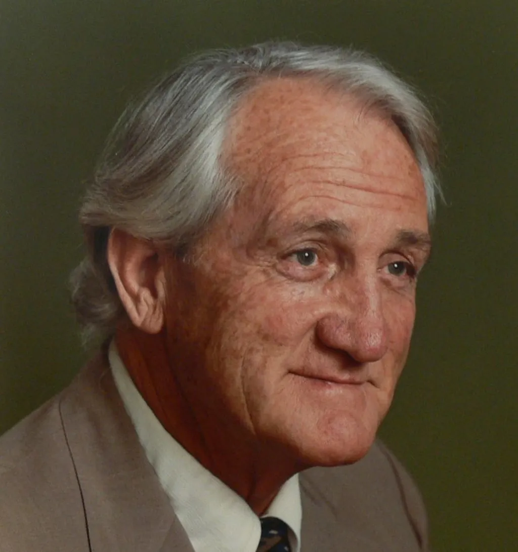 Portrait colour photograph of a man, Tom Uren, smiling and wearing a beige-gray suit and tie.