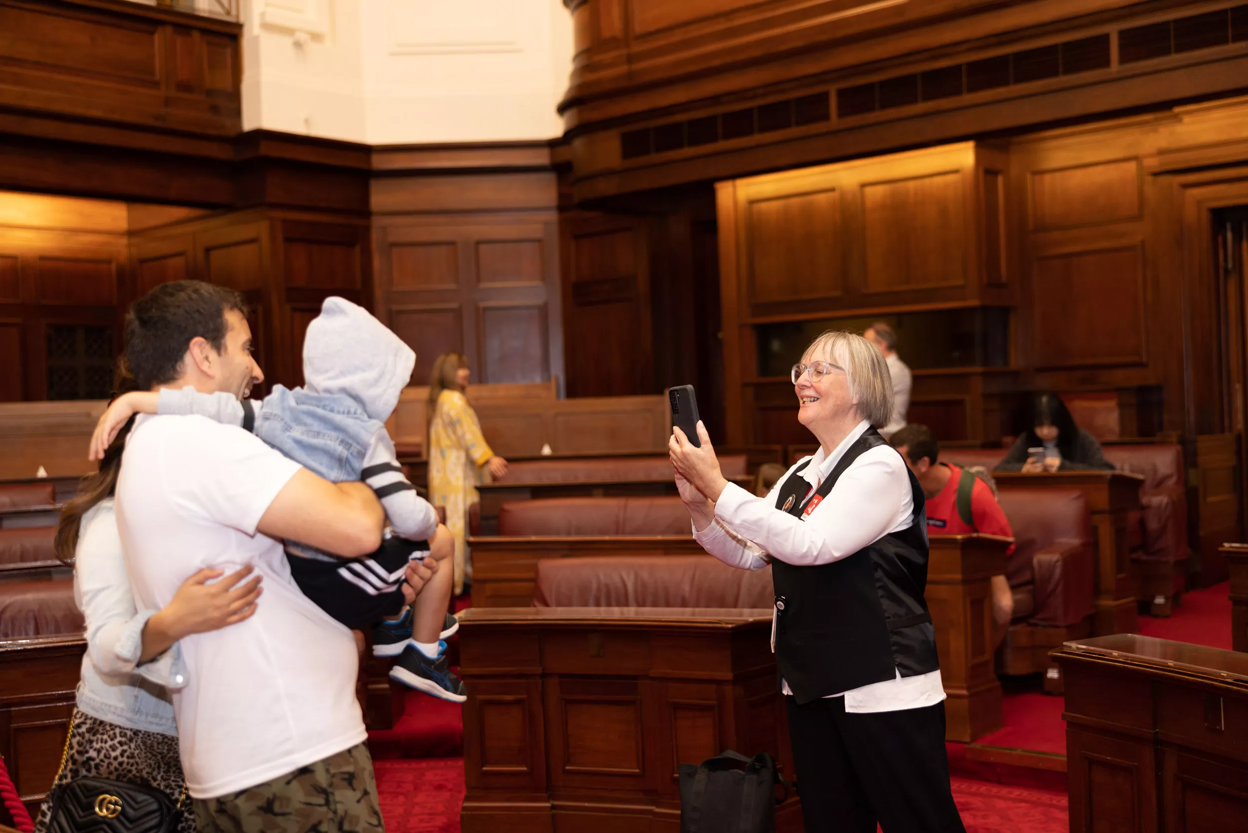 A volunteer takes a photo with a phone of a couple holding a young boy in the Senate Chamber. 