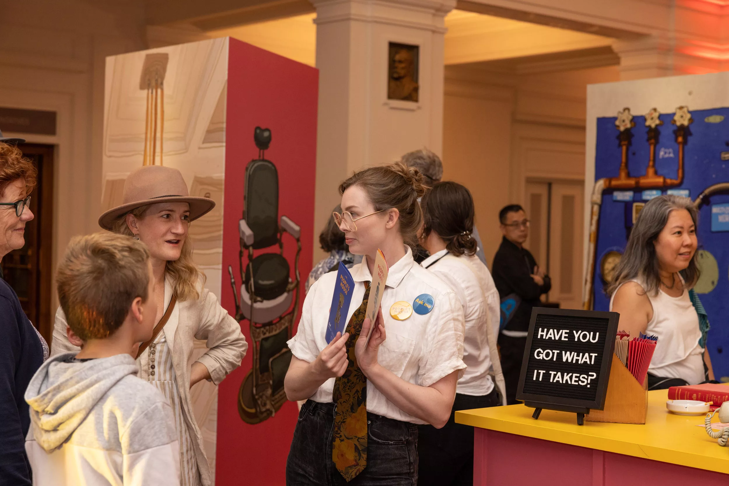 A woman with a white shirt and tie stands in front of a colourful display talking to a woman and young boy and showing them pamphlets.