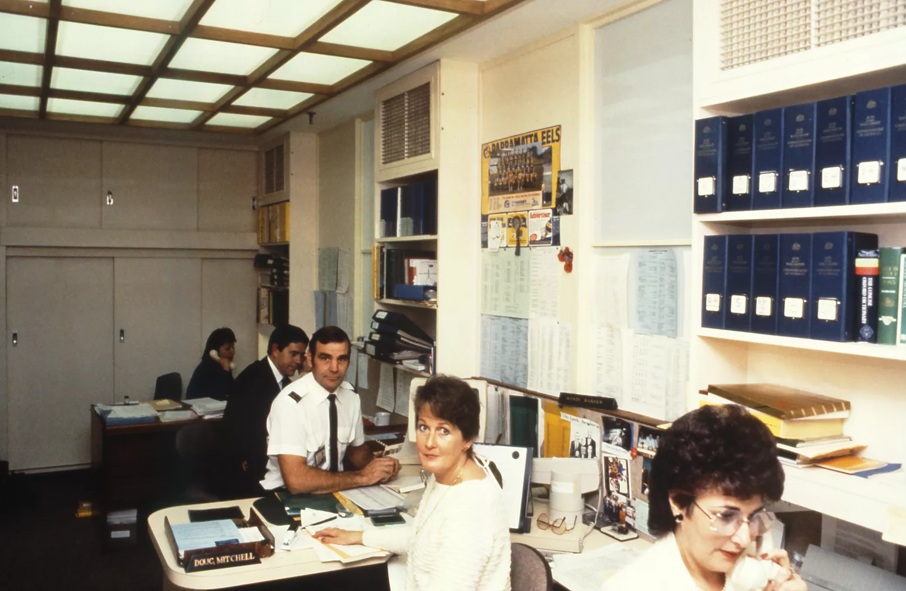This professional colour photograph shows a busy and crowded open plan office in the Speaker’s Suite in 1988 – just a few months before parliament moved to Australian Parliament House. All five desks are occupied with Private Secretary Koula Alexiadis, Principal Private Secretary John Porter, Attendant Doug Mitchell, Assistant Private Secretary Wendy Barnes and Bookings Officer Marie Donnelly. Koula, John and Marie are all on the telephone while Doug and Wendy smile at the camera.