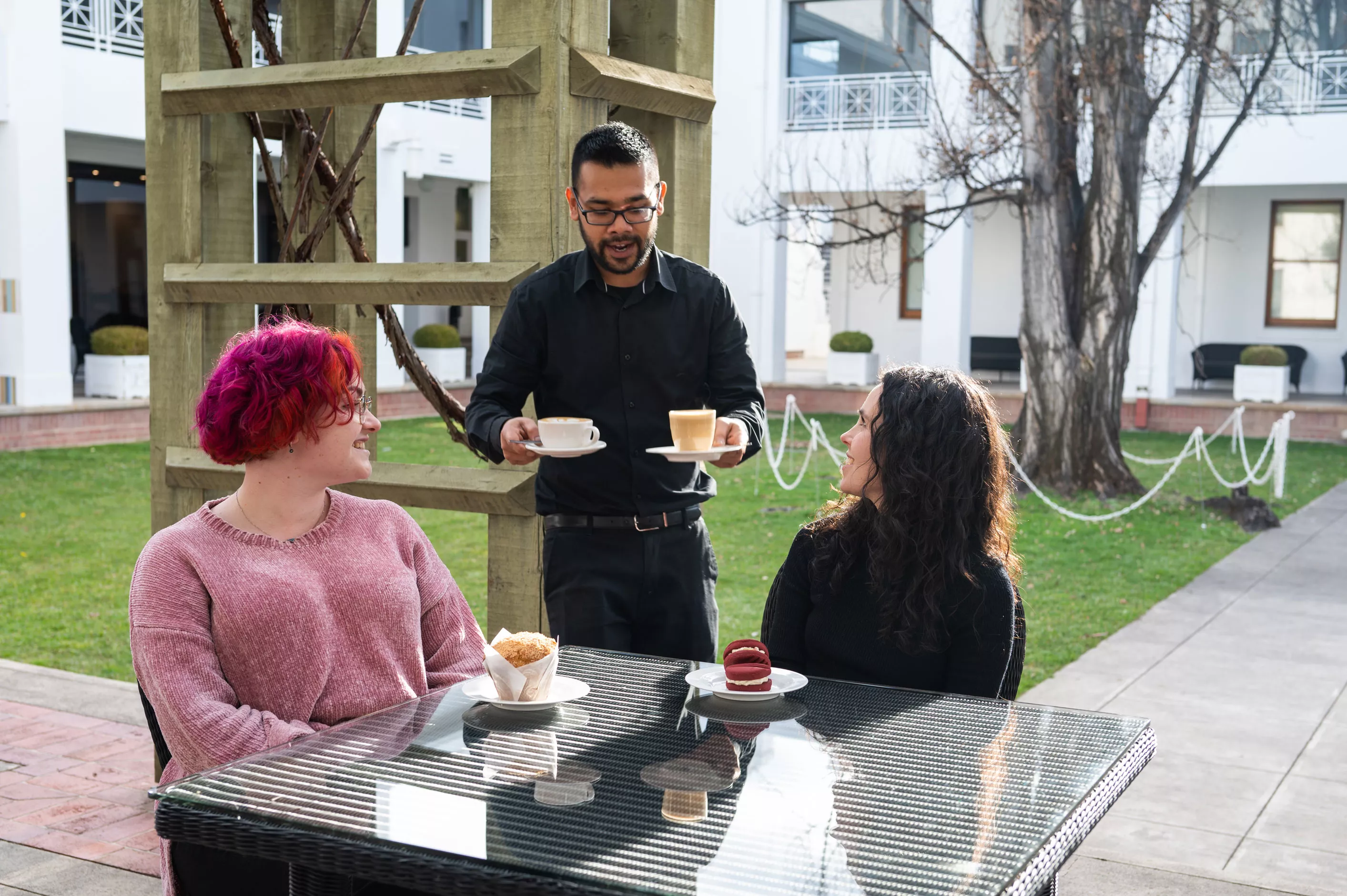 Two women sit at a table in an outdoor courtyard cafe and look up at a waiter bringing them coffee. 