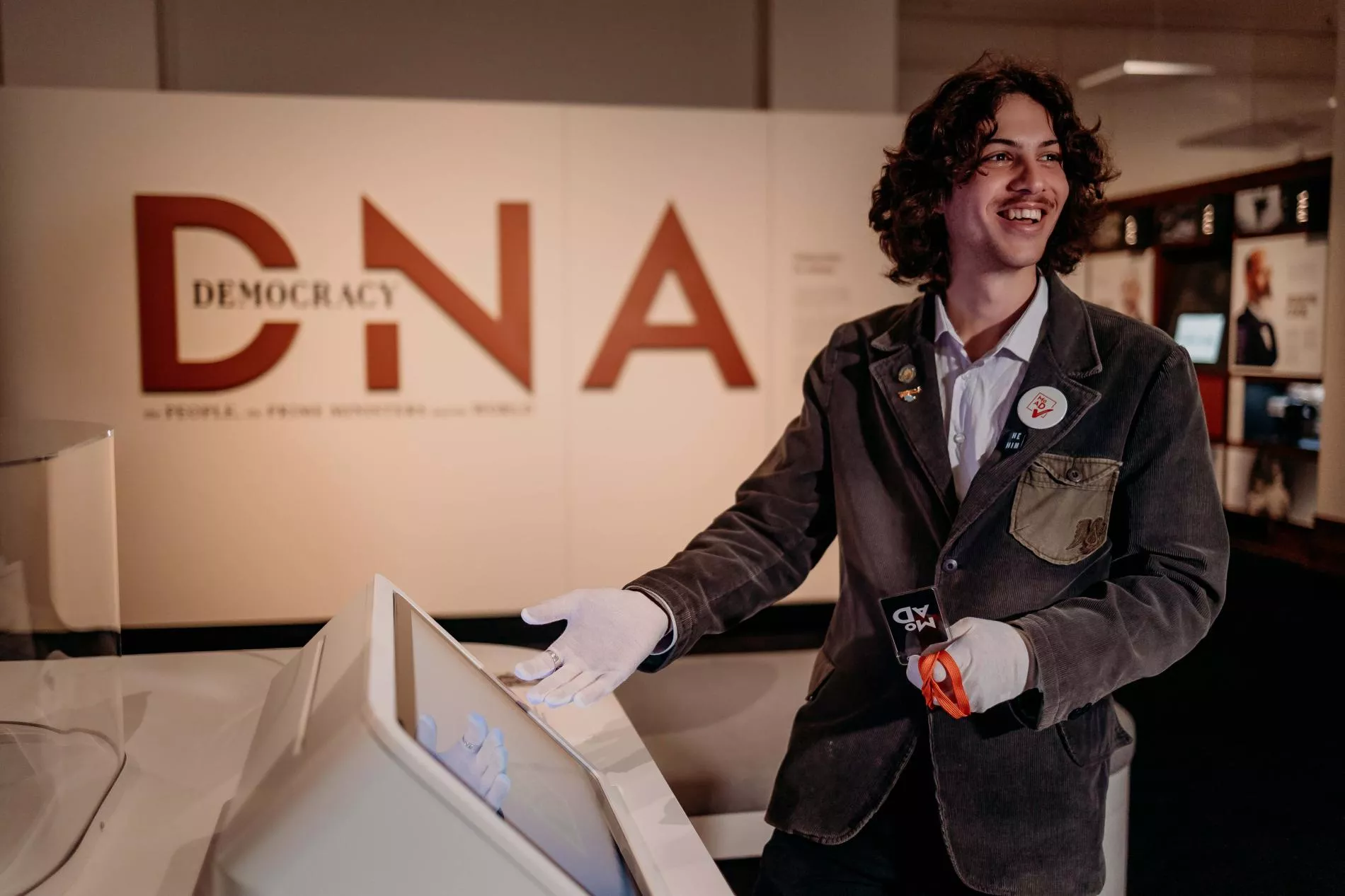 A man in a brown coat and white gloves with a name badge points to a screen. 