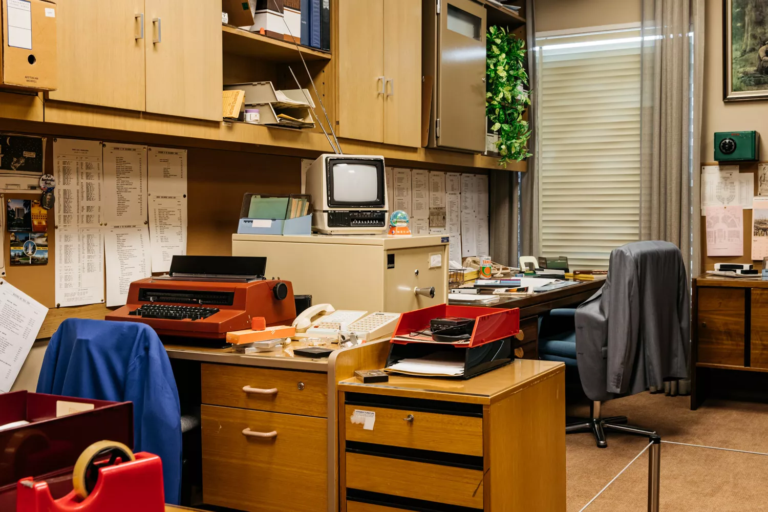 Styled like it's from the '80s, this is the corner of an office typewriters, desks, chairs with jackets over them.