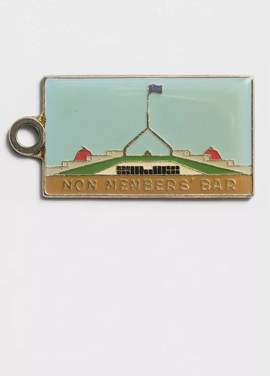 Photograph of a metal tag with an enamel illustration of Provisional Parliament House and the label "Non members bar"