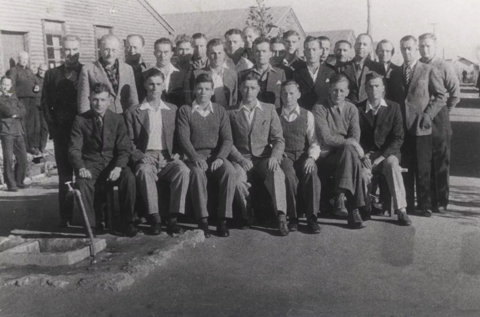 About 25 men in suits or smart casual clothing posing for a group photograph looking dignified towards the camera but not smiling. They are in a military camp in front of long wooden huts.  