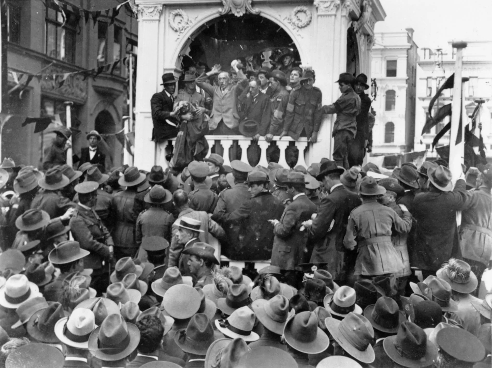 A black and white photo of large group of people wearing hats crowd around a podium where Billy Hughes is giving a speech. A black and white photo of large group of people wearing hats crowd around a podium where Billy Hughes is giving a speech.