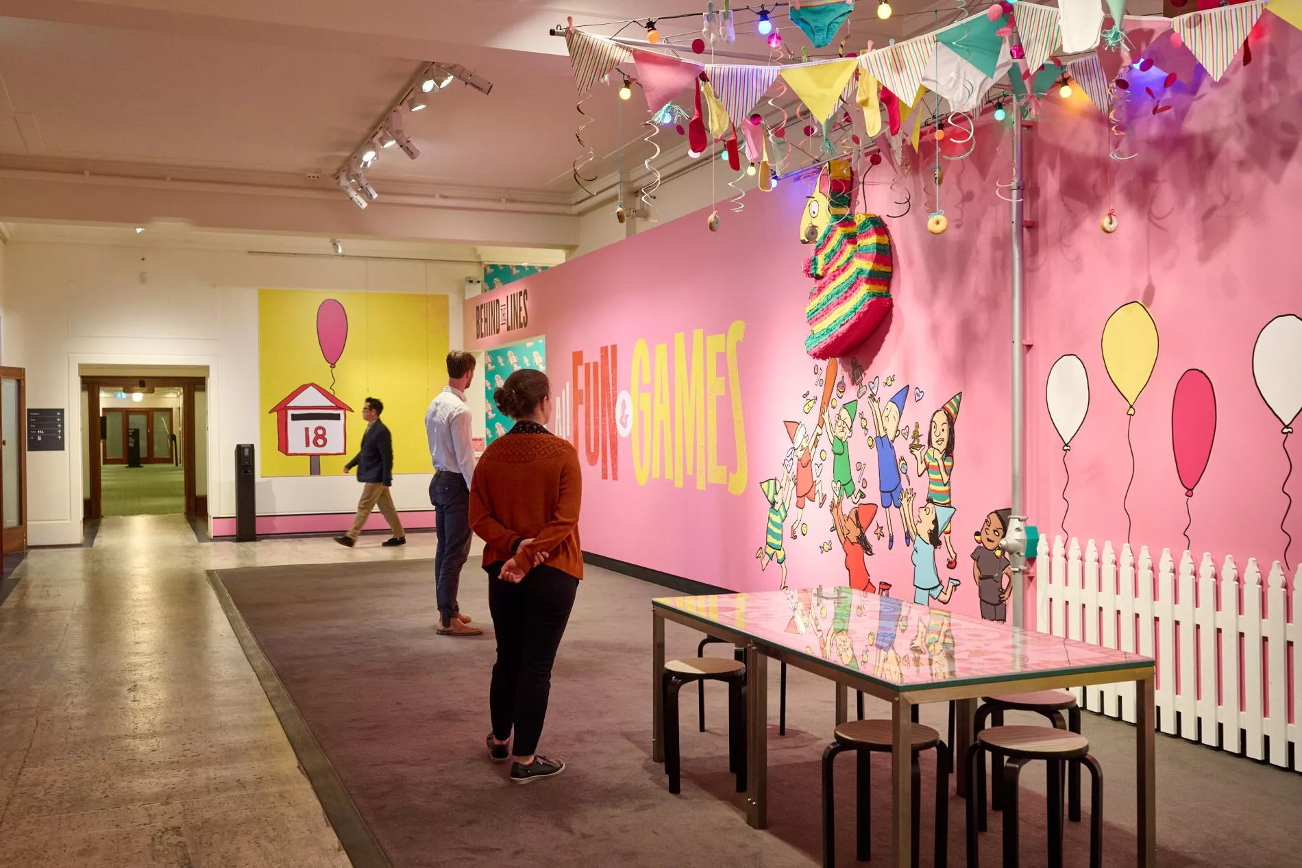 Visitors look at a long musk-pink wall with the words ‘Behind the Lines’ and ‘All Fun and Games’ on the left. On the right is a hills hoist is embedded in the wall and decorated with bunting and streamers. 