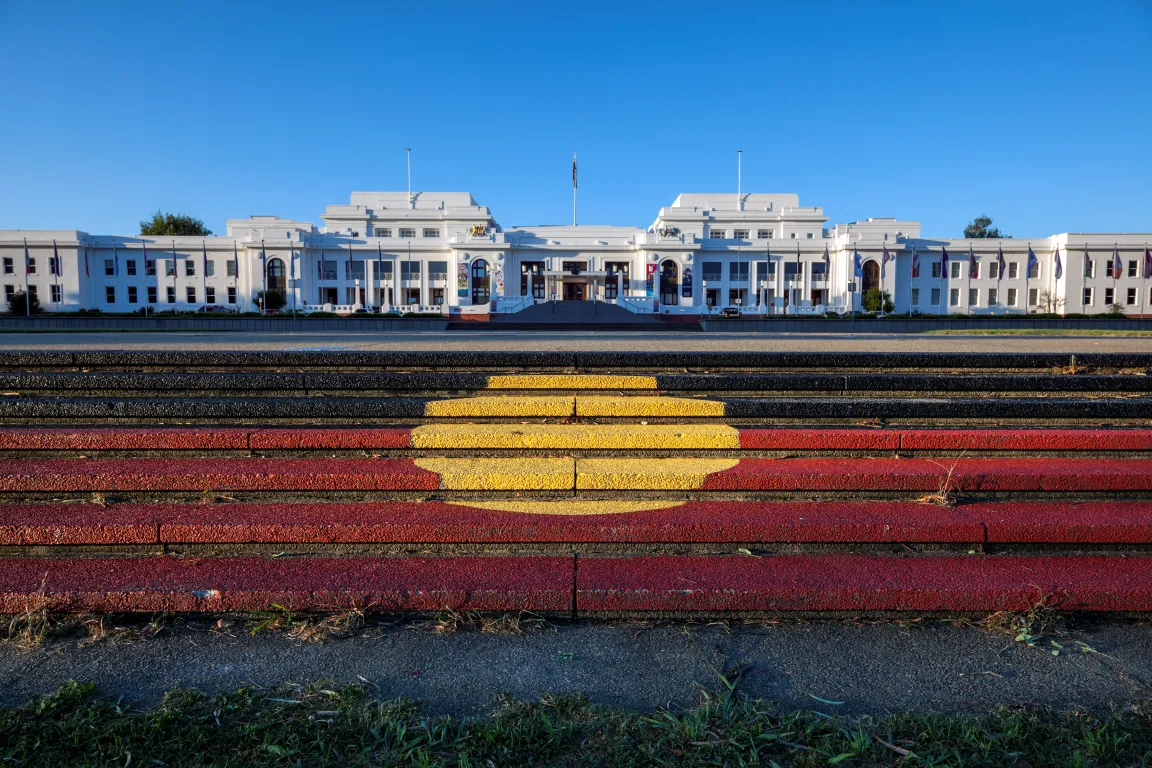 Steps with the Aboriginal flag painted on them lead up to a view of a large long white building, Old Parliament House.