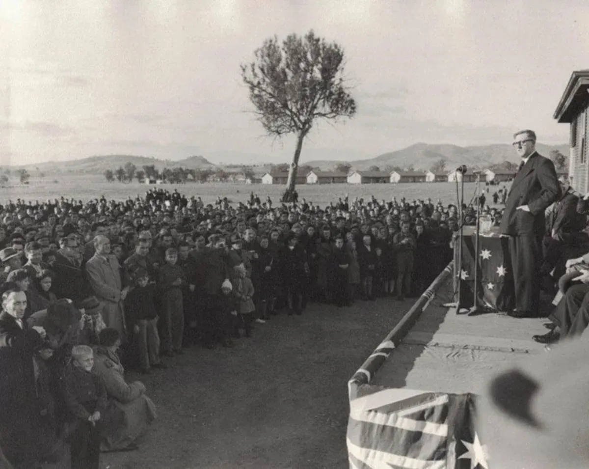 Arthur Calwell stands on a platform with microphones, addressing a crowd of people. In the distance is a row of long buildings.