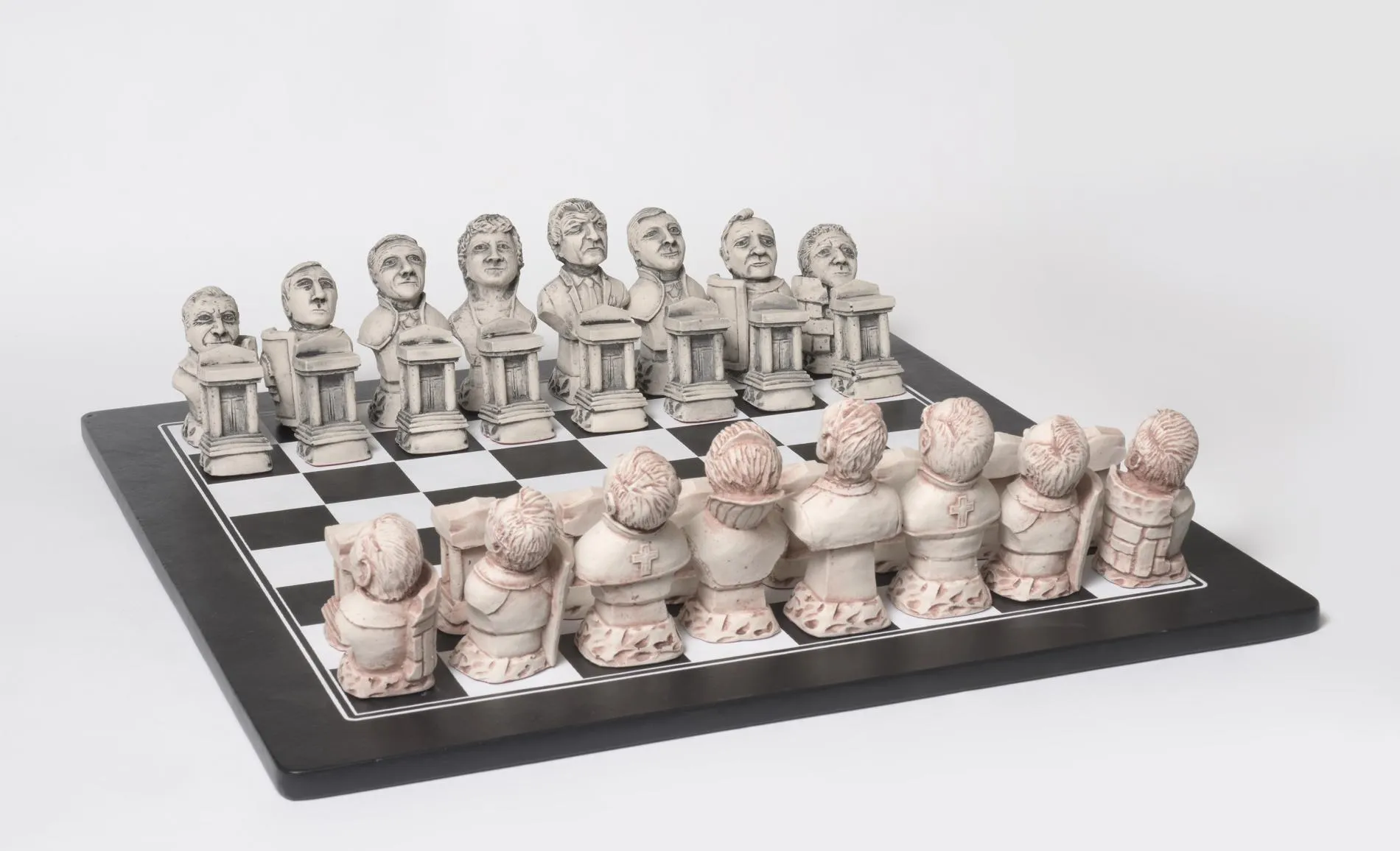 A chess set featuring politicians carved from a stone. The faces of the black chess pieces are visible and the backs of the white pieces. 