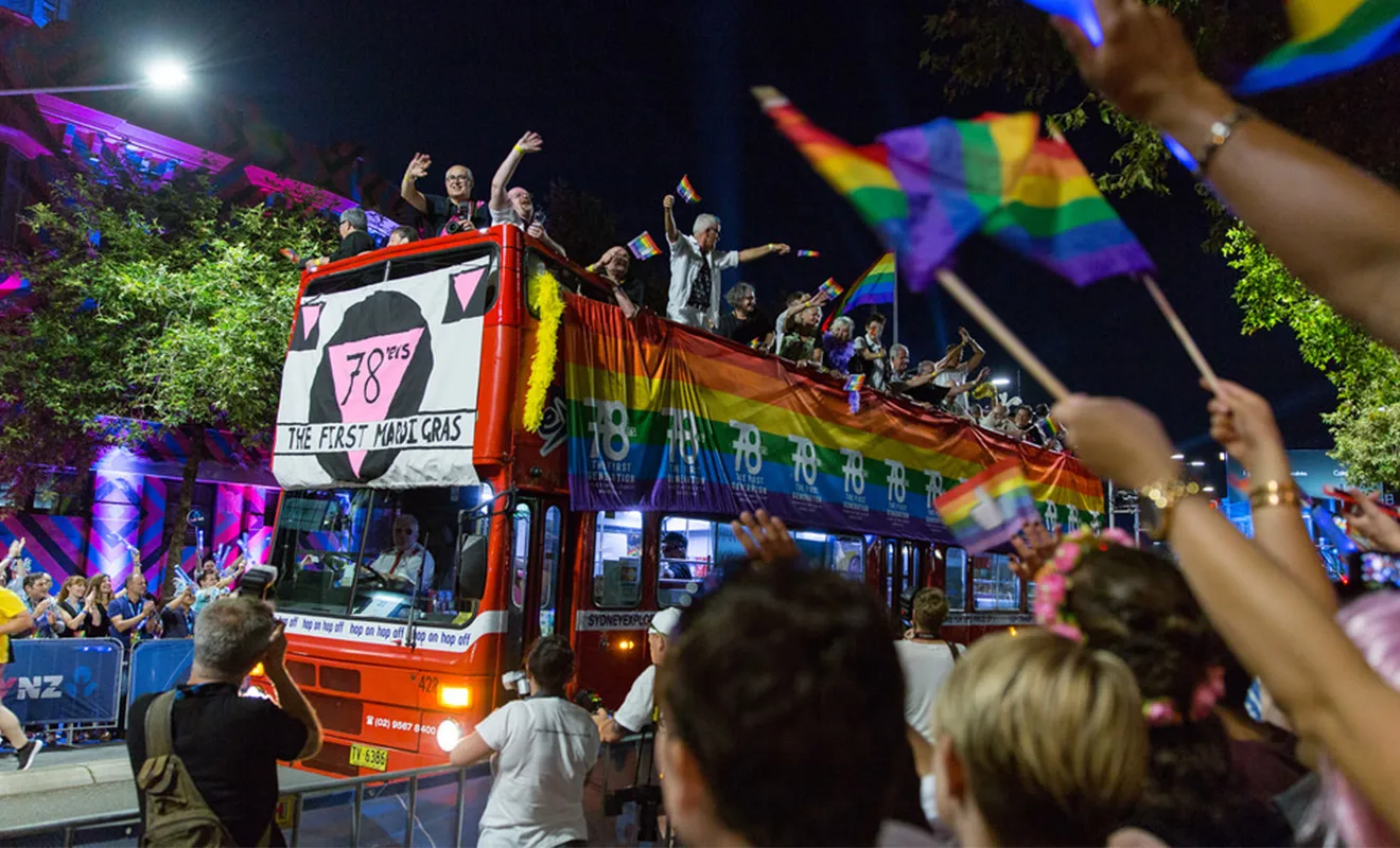 A red double-decker bus with rainbow banners displaying the words '78ers: the first mardi gras' with people on top of the bus waving to crowds on each side of the street.