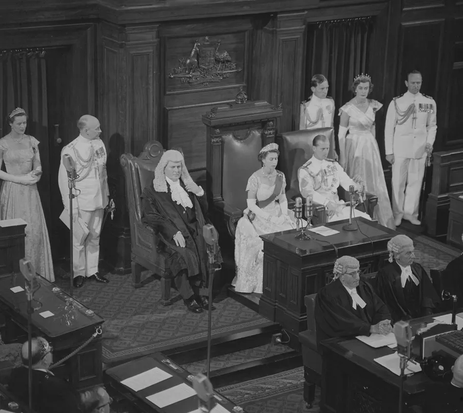 Queen Elizabeth II, with Prince Phillip, sitting in the Senate Chamber opening parliament. Various aides and parliamentary officials surround them.