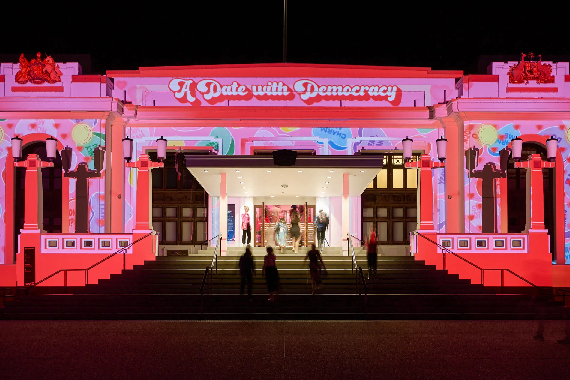 Pink and red illuminates the front of Old Parliament House. Text in white and red above the entrance reads 'A date with democracy'. Pink and red illuminates the front of Old Parliament House. Text in white and red above the entrance reads 'A date with democracy'.