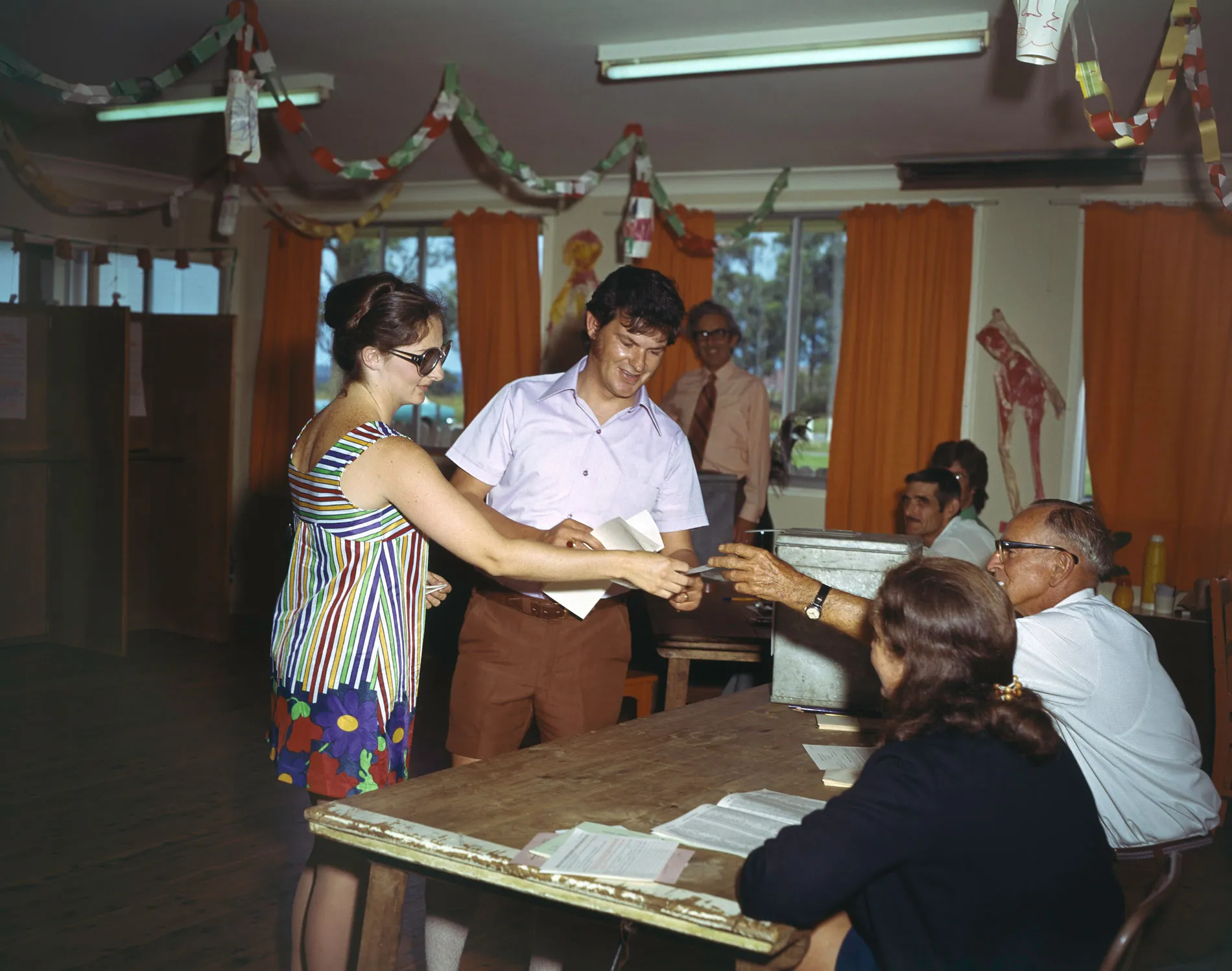 Two people being handed information at a polling booth in a hall with orange curtains. Two people being handed information at a polling booth in a hall with orange curtains.