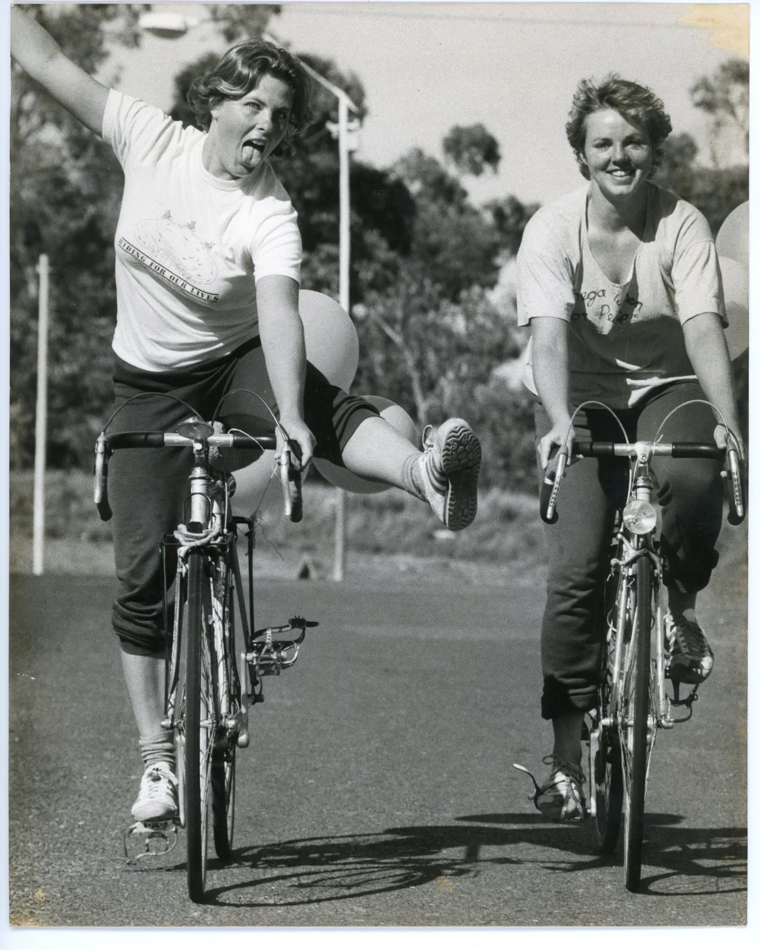 Two women ride bicycles, one woman has her arm raised and the opposite leg kicking up from the bike pedal. Two women ride bicycles, one woman has her arm raised and the opposite leg kicking up from the bike pedal.