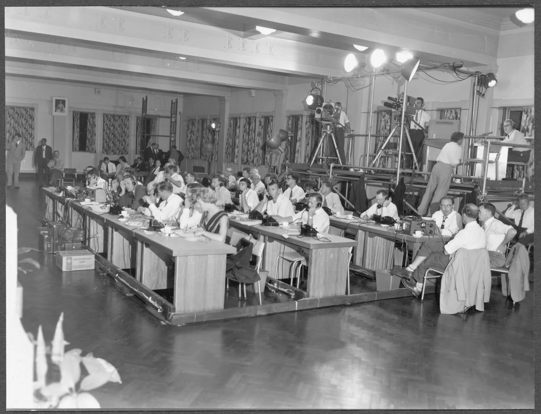 A room full of people at desks on telephones, behind them is a platform with people operating video cameras with some bright lights