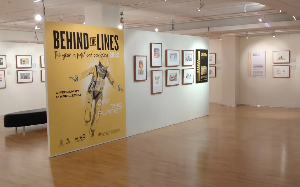 A gallery space with framed cartoons on the walls. A large, yellow introductory panel features the illustration of an astronaut and the words 'Behind the Lines'.