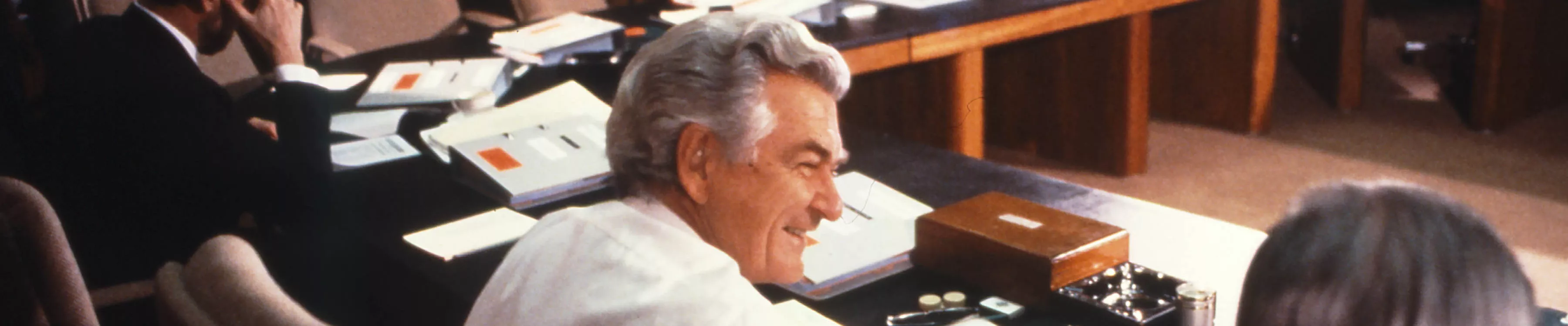 This colour photograph is taken from behind Deputy Prime Minister Lionel Bowen who is seated beside Prime Minister Bob Hawke. Both are dressed in standard business attire.  Other members of Cabinet can be seen sitting around the table although only half of the seats are filled. In the background are a photographer and two cameramen photographing and filming the cabinet meeting. 