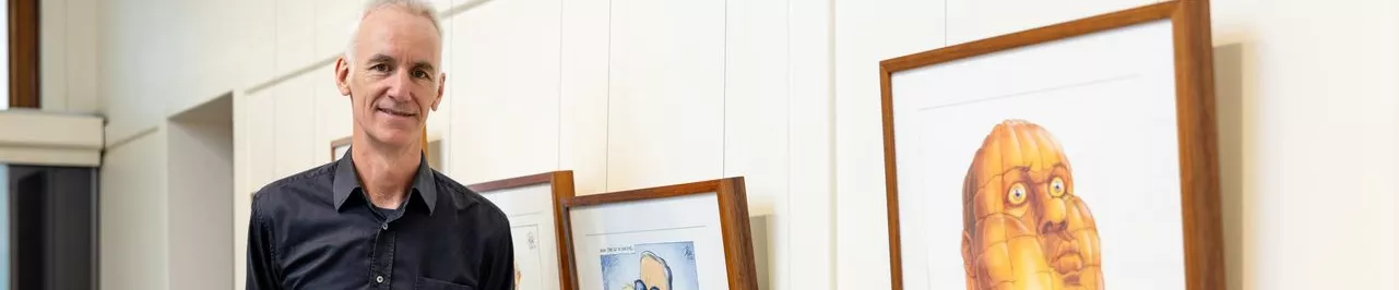 David pope stands next to a framed print of one of his cartoons