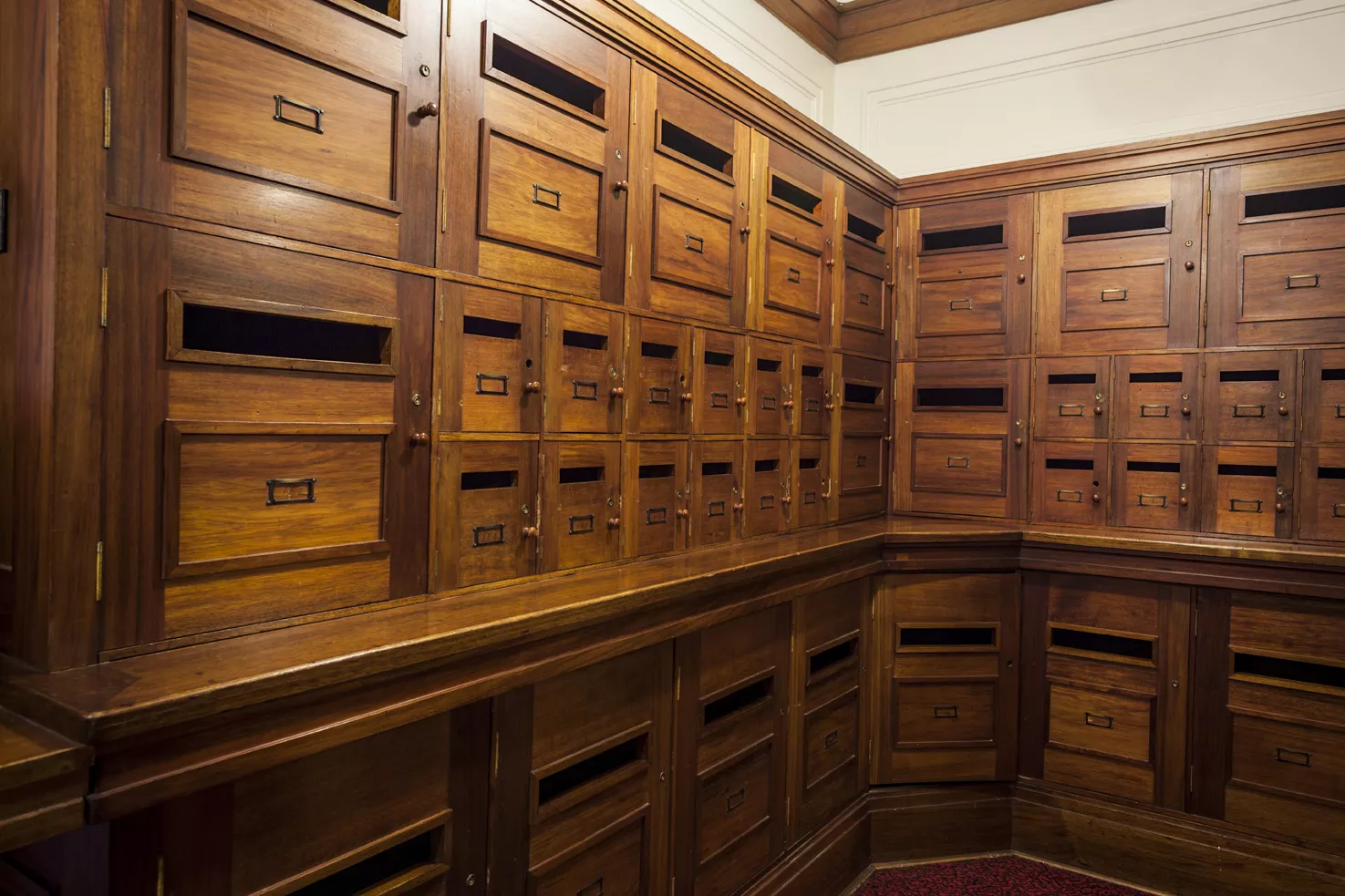 Many wooden pigeonholes with doors, wrapping around the corner of a room. 