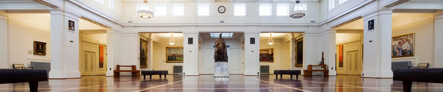 Photograph of a large room with wooden flooring, white walls and ceiling, and a statue of King Geroge V at the centre back.  