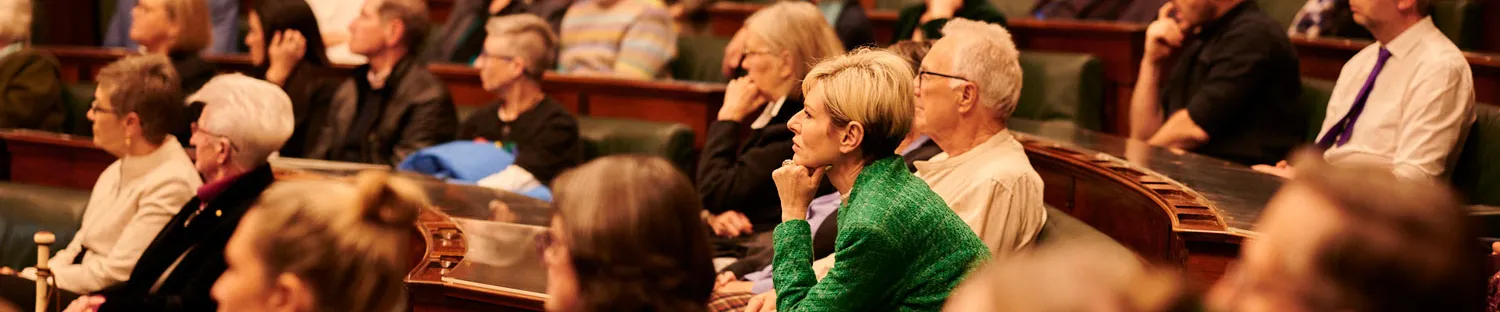 An audience sits on green leather benches in the House of Representatives at Old Parliament House. The room has wood panelled walls.